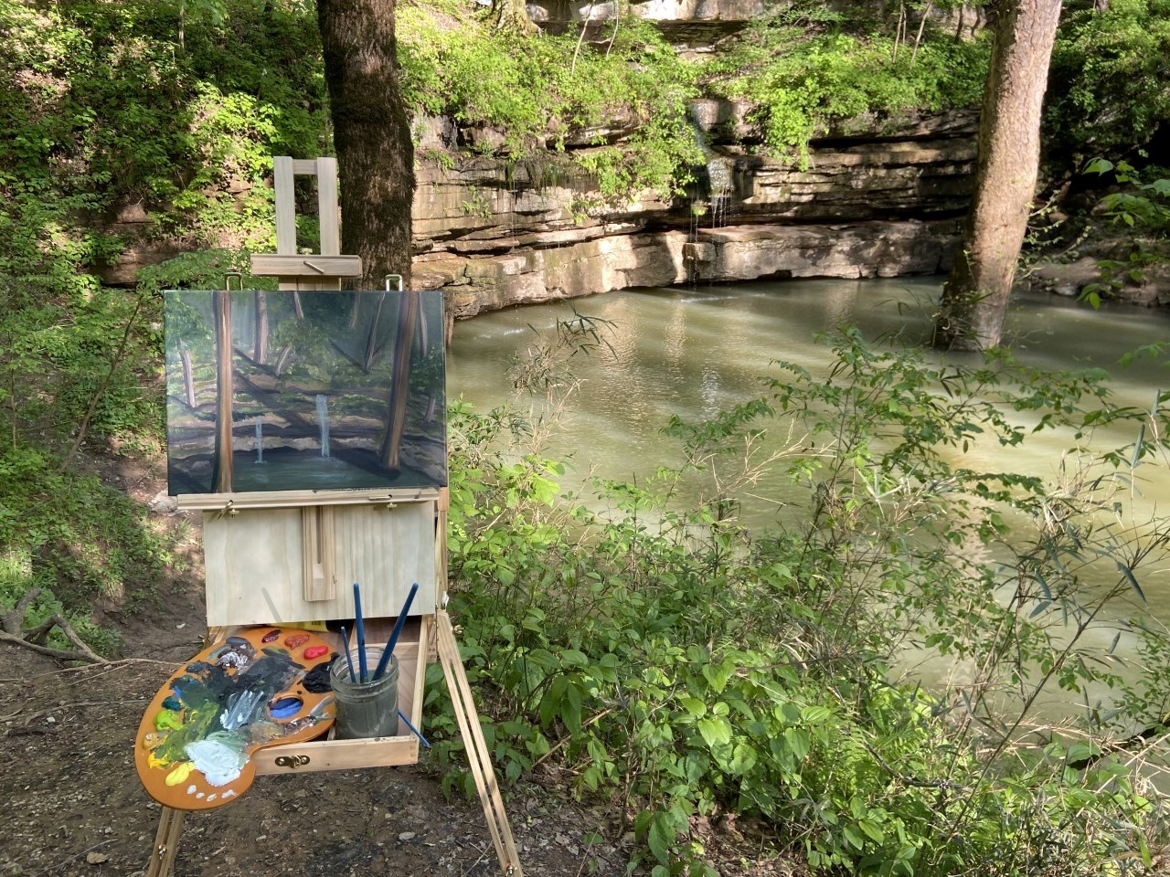 An artist easel displays a painting of a body of water in a forest area while the same body of water and forest is displayed in the background.