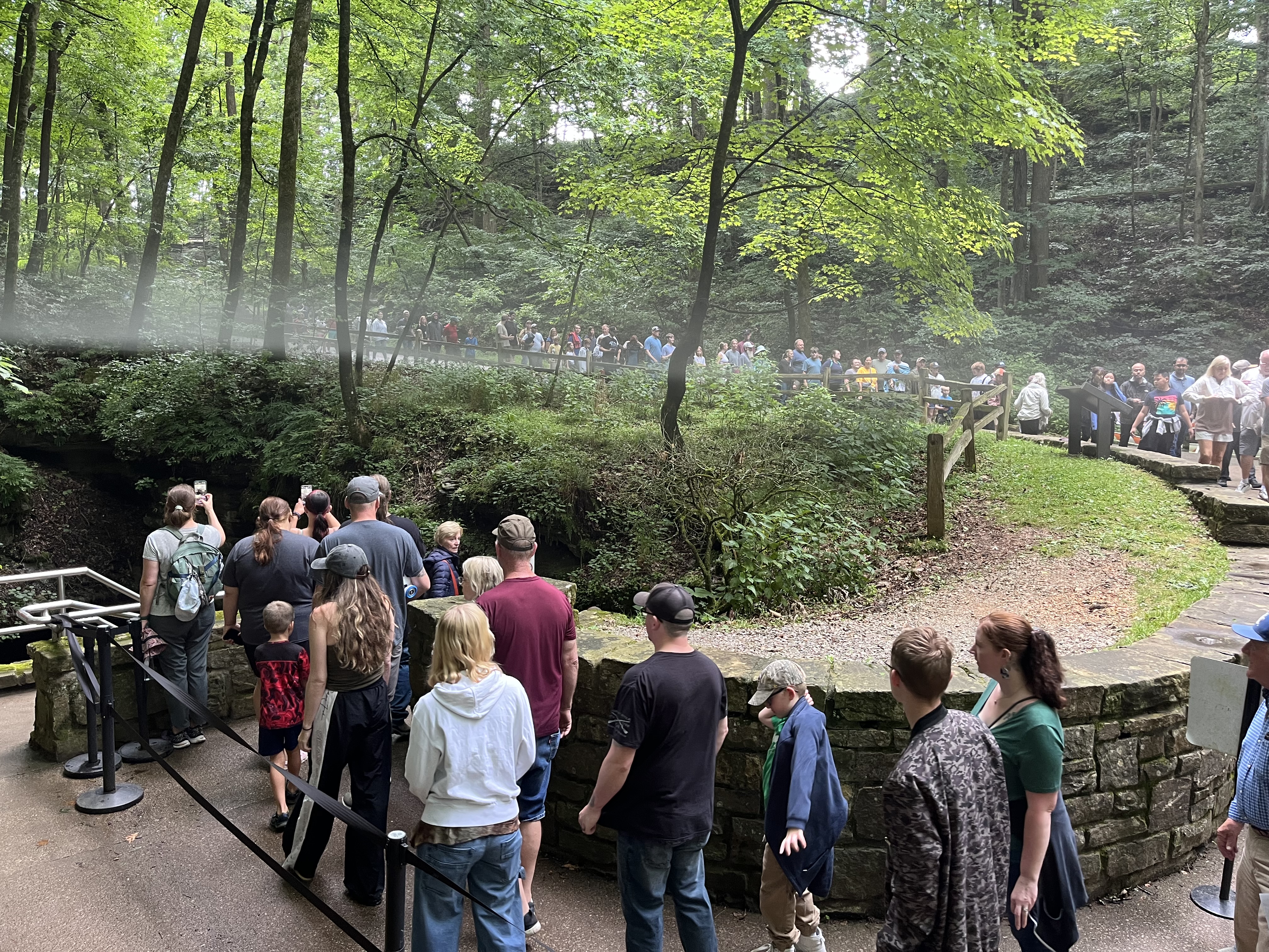 A large group walks down a curved pathway toward a cave entrance in a wooded area.