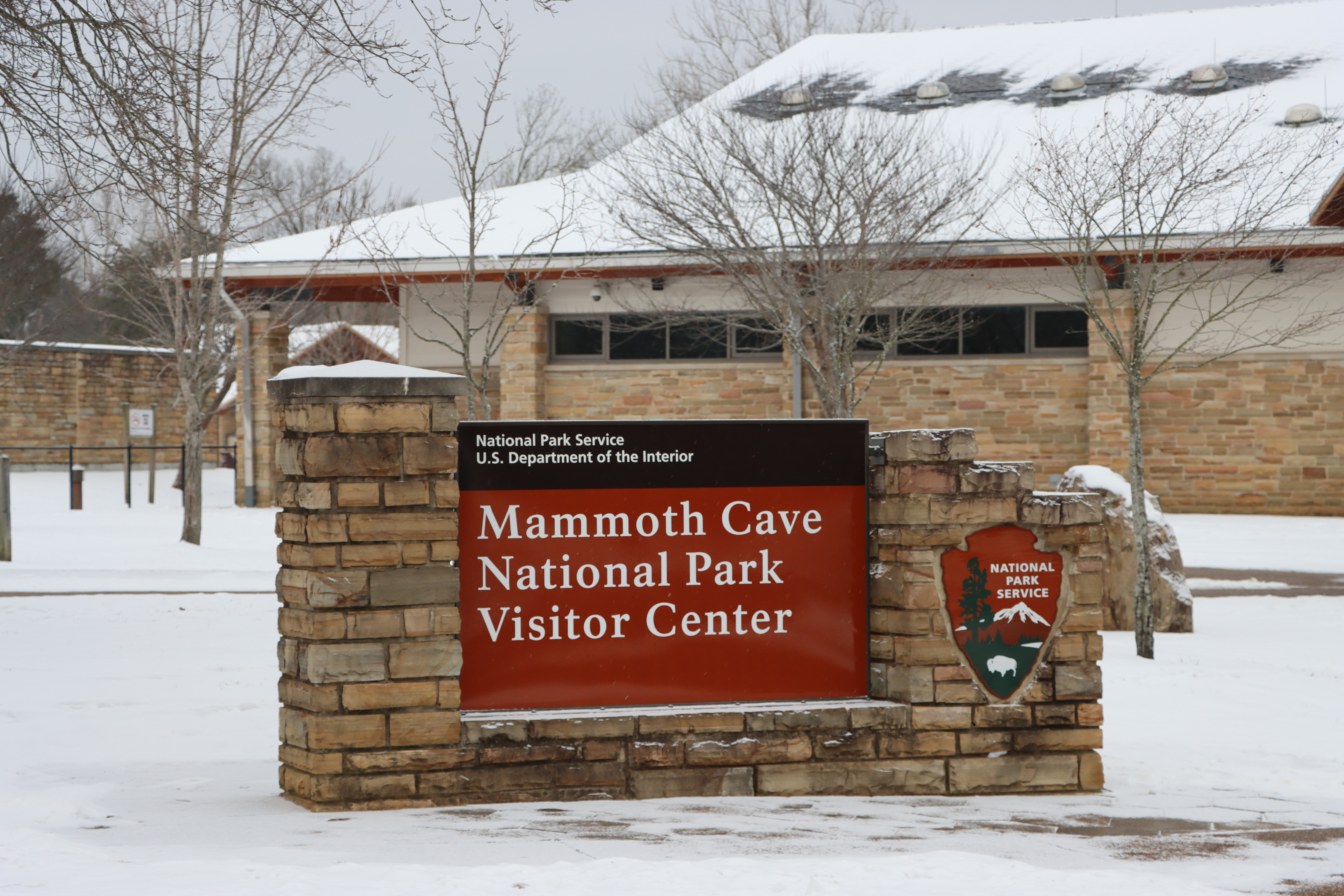 A sign with the NPS Arrowhead and "Mammoth Cave National Park Visitor Center" surrounded by snow.
