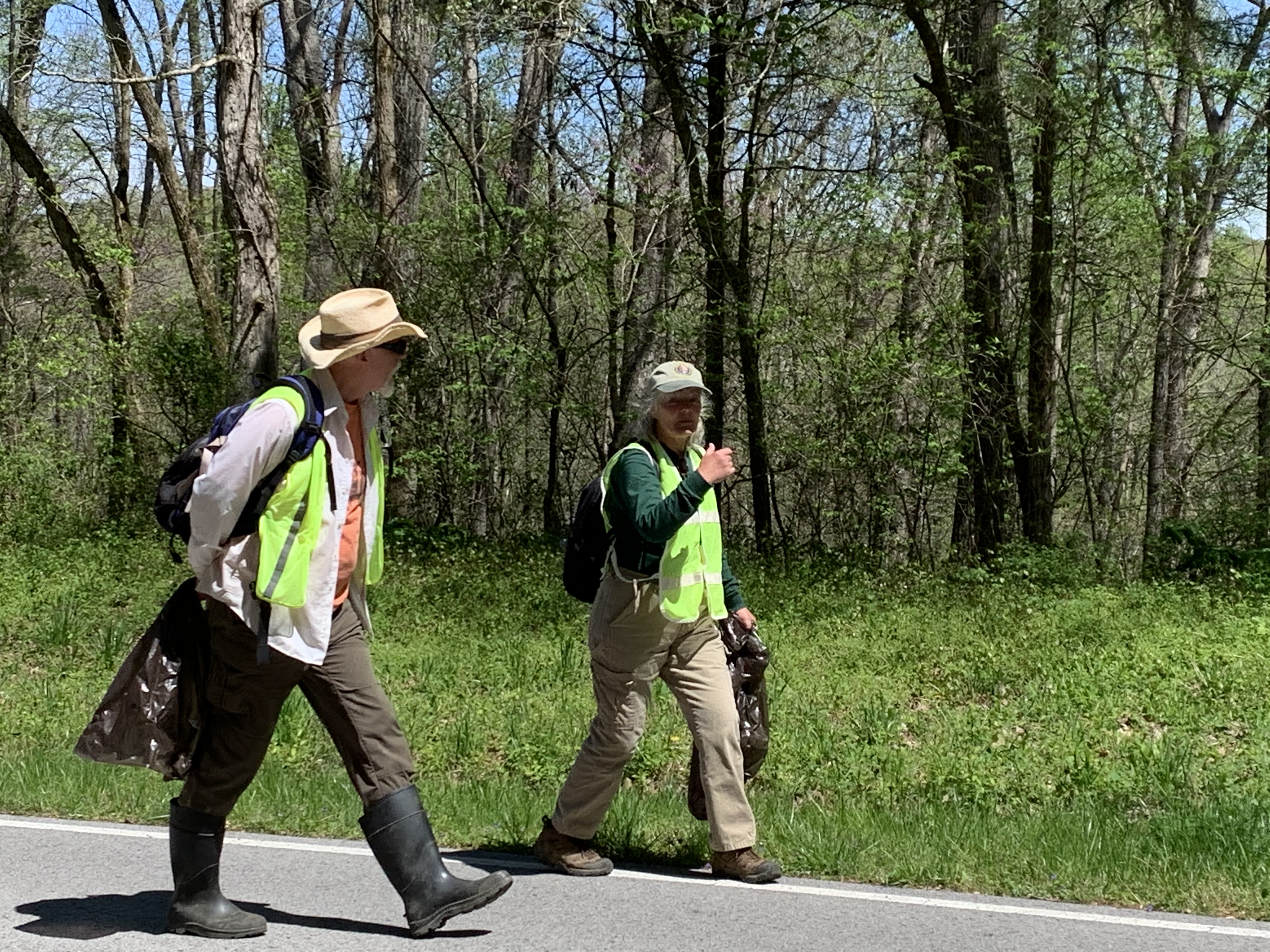 Two people carrying trash bags walk along a roadside.