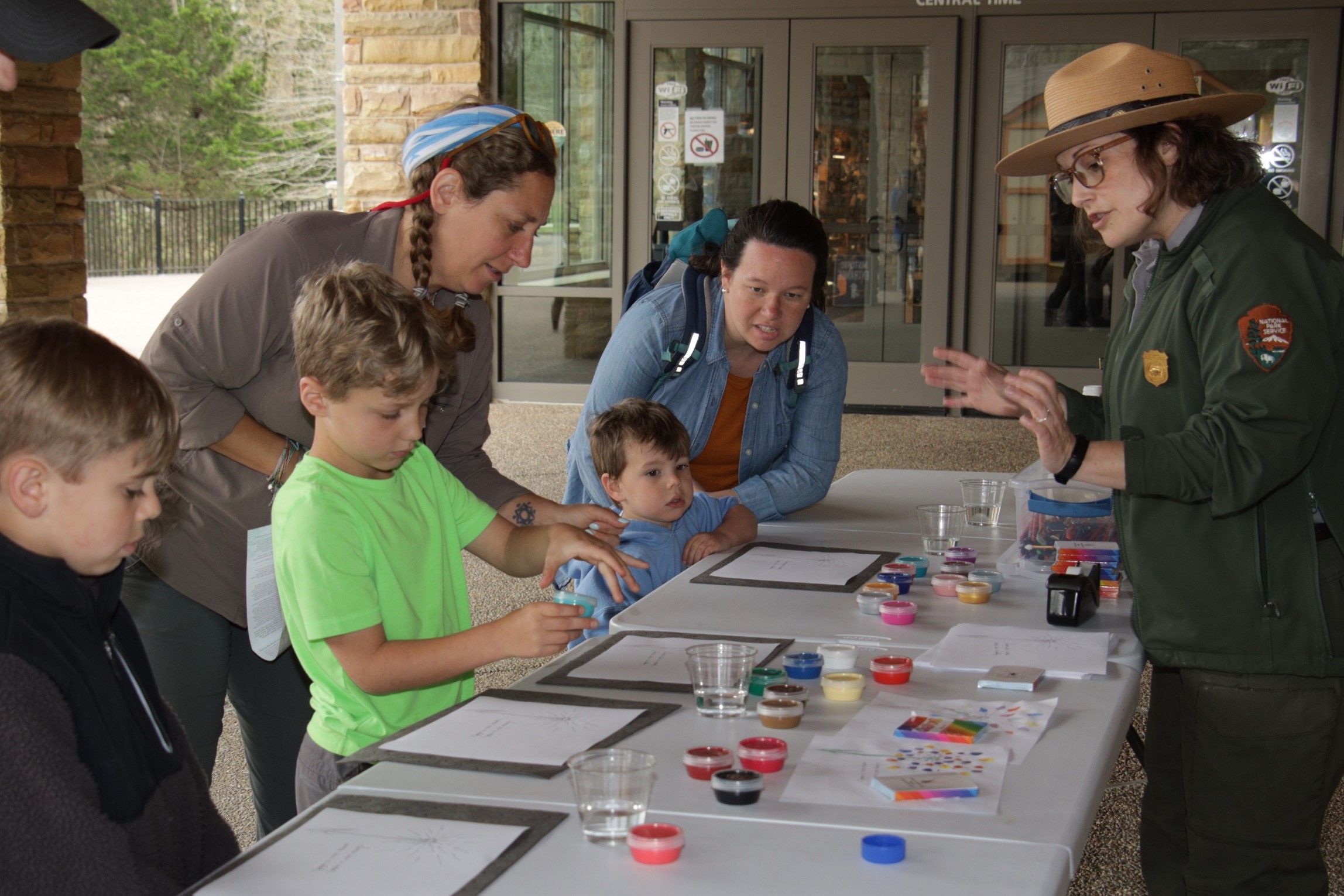 Five people stand next to a table with paper and paints while a woman in a green park service uniform talks to them