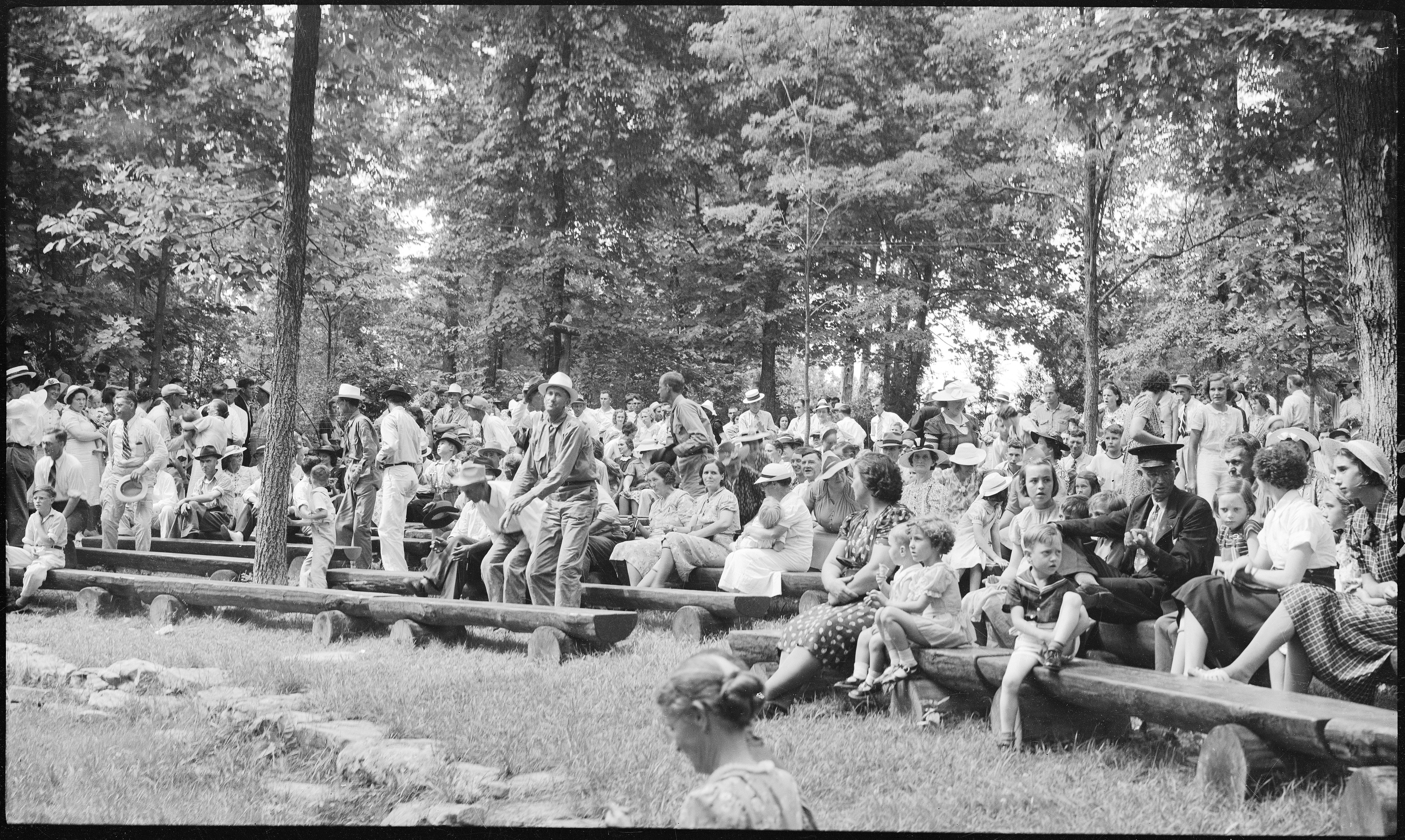 A historical photo of a group of people gathered in an outdoor amphitheater with wooden benches.