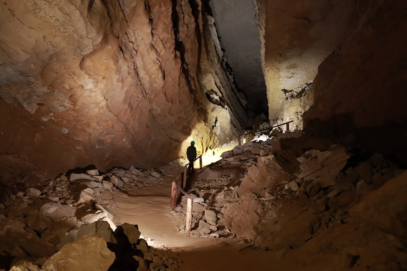 A person walks up a steep hill in a dimly lit cave passageway with tall ceilings.