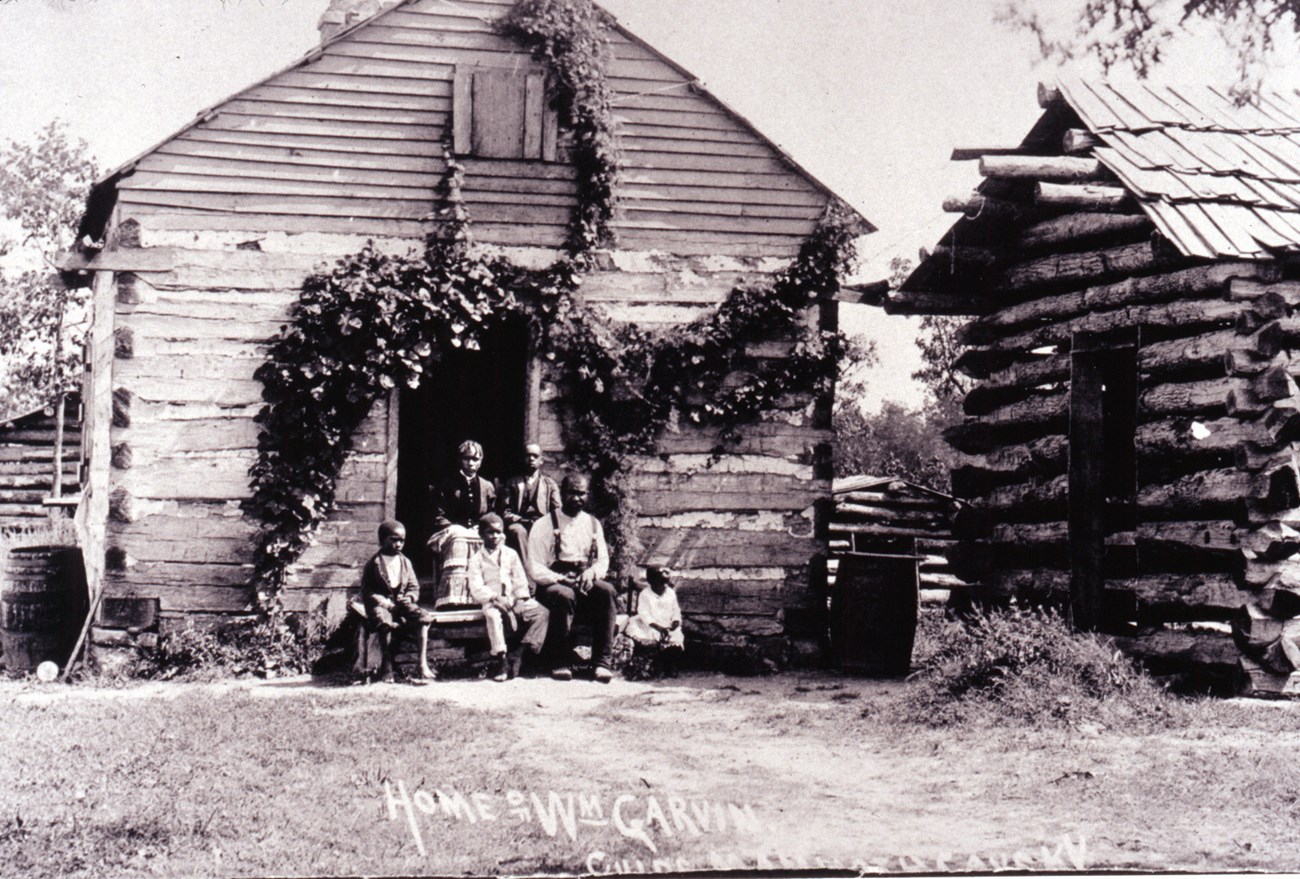 Black and white photo showing a wooden cabin with family of six people sitting on the front doorstoop.