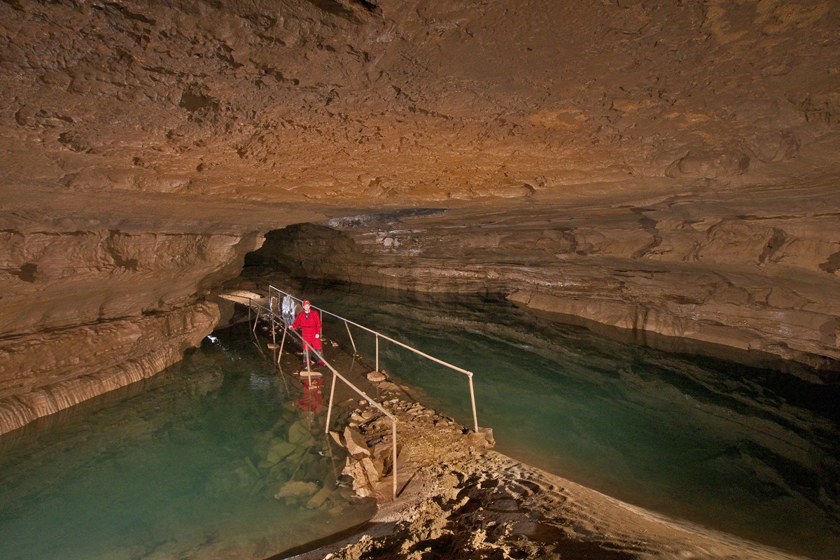 A man wearing red coveralls standing in a pool of water in a large cave passage.