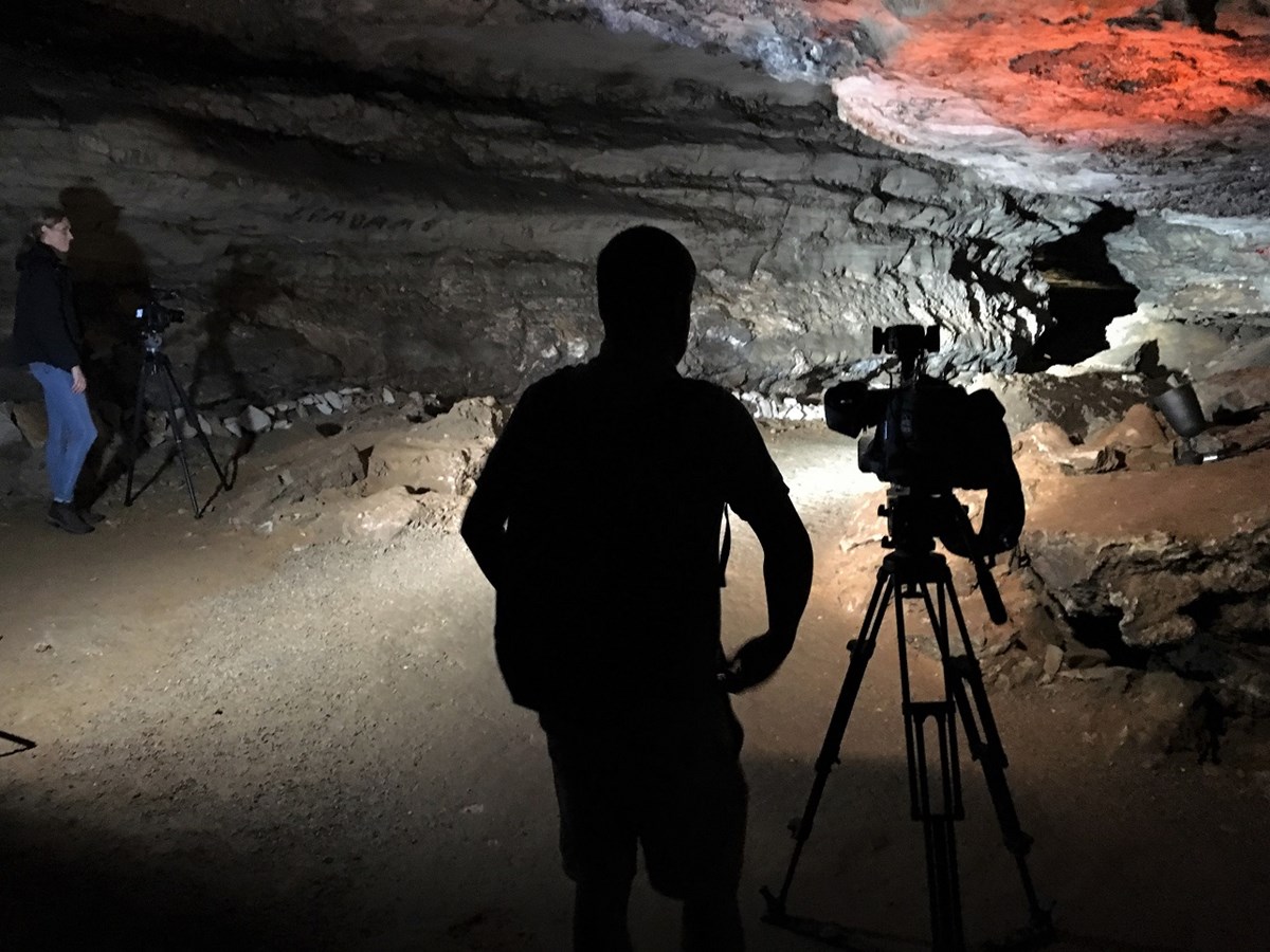 A person and film camera are silhouetted against a rocky cave backdrop