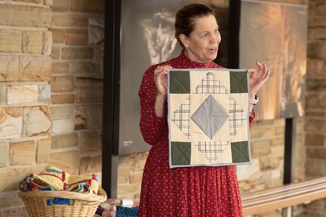 A woman holds up a square piece of quilted cloth that shows a pattern of squares and triangles.