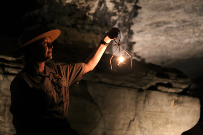 A ranger standing inside the dark interior of Mammoth Cave holds up a historic caving lantern.