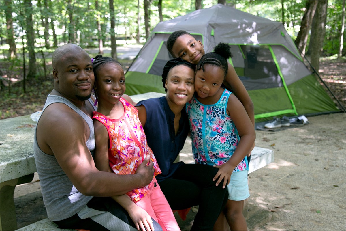 A family, with a father, mother, two young daughters and a young son near their tent. All are smiling and looking toward the viewer. A family, with a father, mother, two young daughters and a young son near their tent. All are smiling and looking toward the viewer.