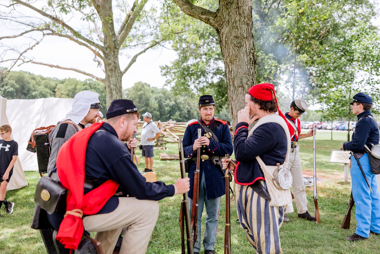 A group of men in various uniforms from the Civil War stand around and chat, some in front, some on the periphery. In addition, tents, smoke frok a camp fire, and a couple of visitors look on.