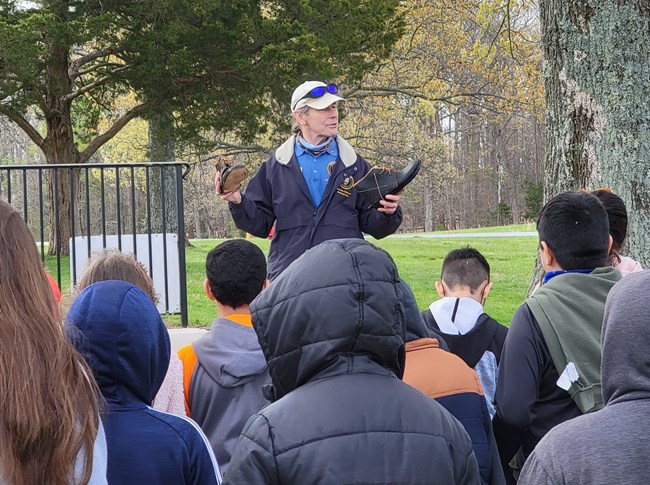 A park volunteer demonstrates the shoes worn by soldiers to students on a chilly morning outdoors.