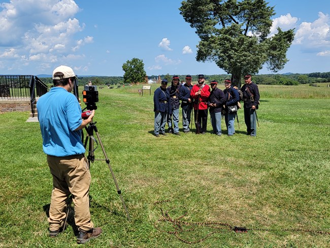 An intern offering a photography program is shooting a photo of living historians in a field during the First Battle Anniversary on a sunny day in July.