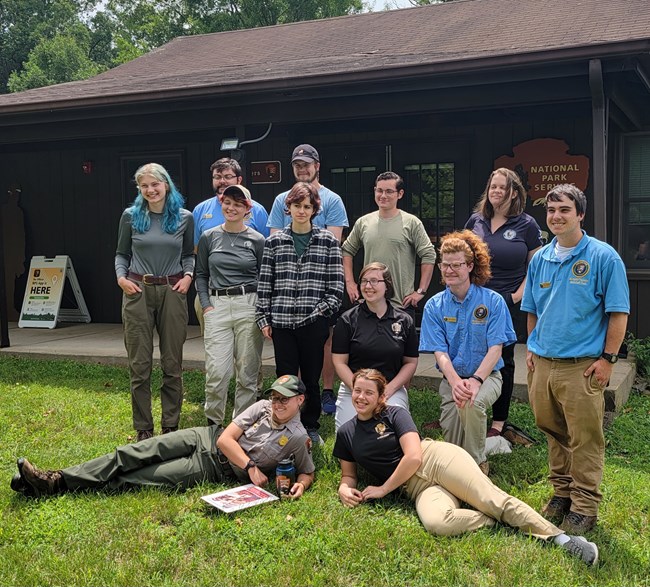 A group of interns in various uniforms sit/lay/stand while posing for a picture outside of the park headquarters in July 2023.