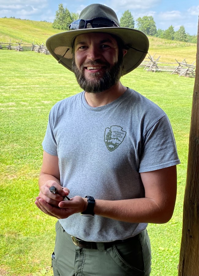 A man in a gray tee and green pants wearing a work hat stands in semi-profile holding somethingi n his hand.