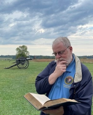 Color photo of a park volunteer reading from a book outside. He has a thoughtful look on his face. Behind him is a grassy field with trees and a black Civil War cannon.