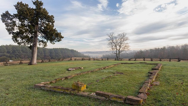 Color photo of the Robinson House site. There are stones outlining the perimeter of where the house used to be and a split rail fence around the property. There are two trees in the image, and the ground is covered with grass.