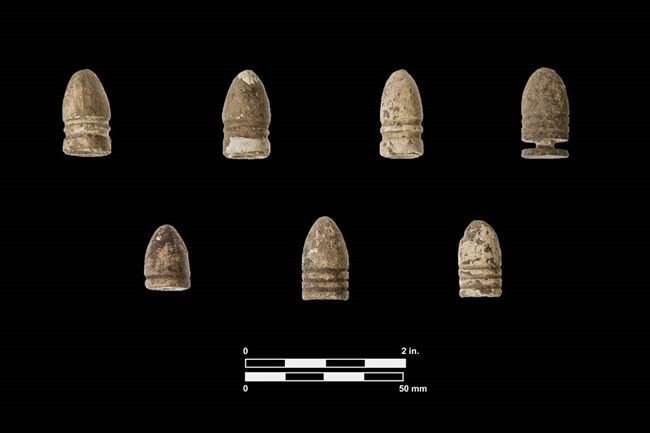 Color photo of seven bullets on a black background. Some of the bullets are similar, but none are exactly the same. All area light brown color and are coated with old dirt but have been cleaned to see detail.