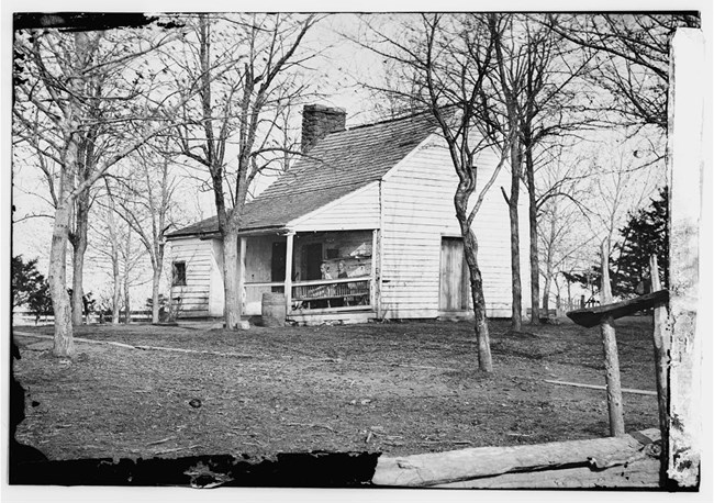 A black and white image of a small white house with a porch. Two women stand/sit on the porch.