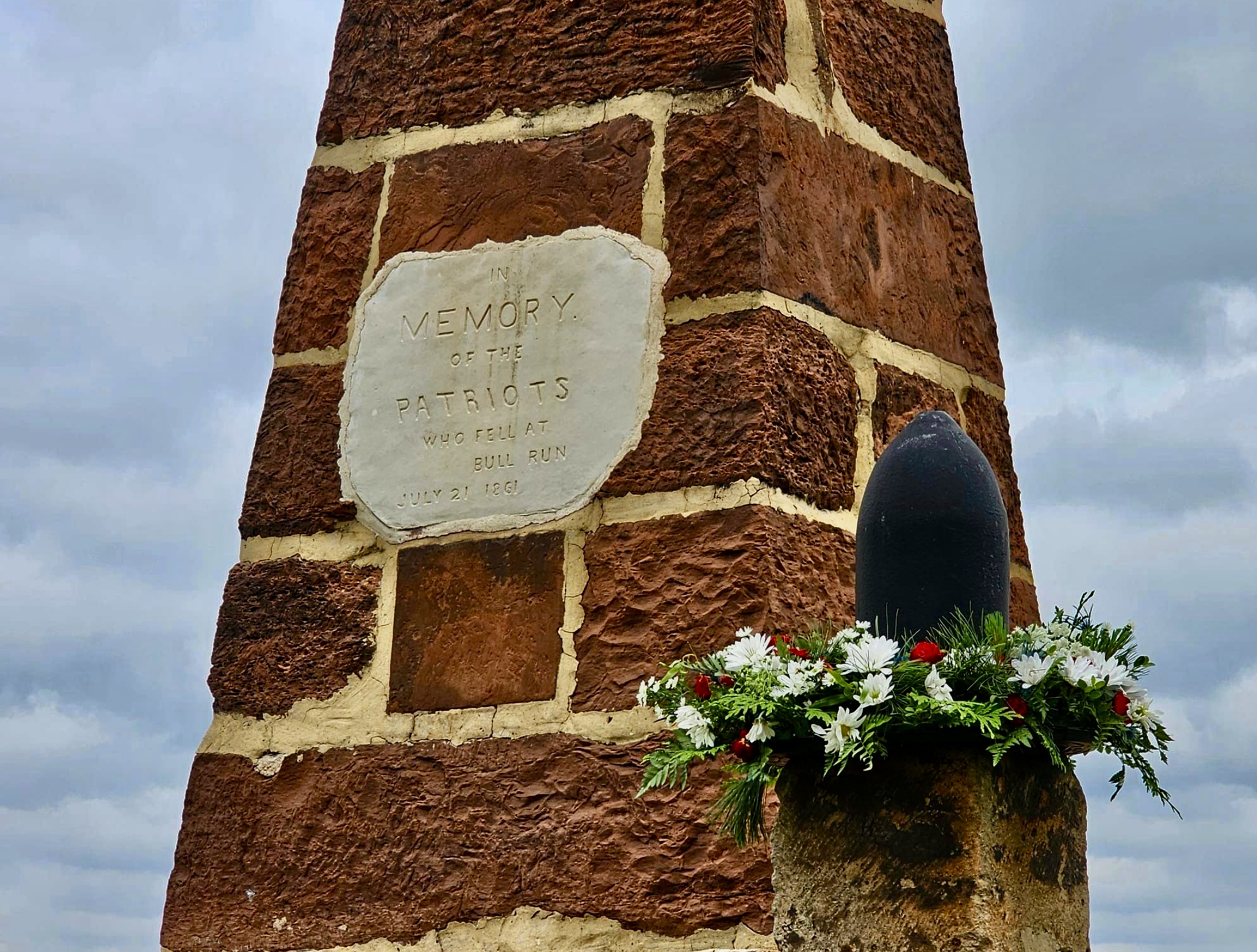 Photograph of the Patriots Monument with flowers around one of the large black iron shells.