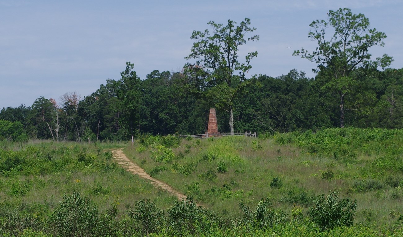 The Deep Cut with the Groveton Monument: The terra cotta colored obelisk sits on the crown of a hill lined with green brush and trees in the background.