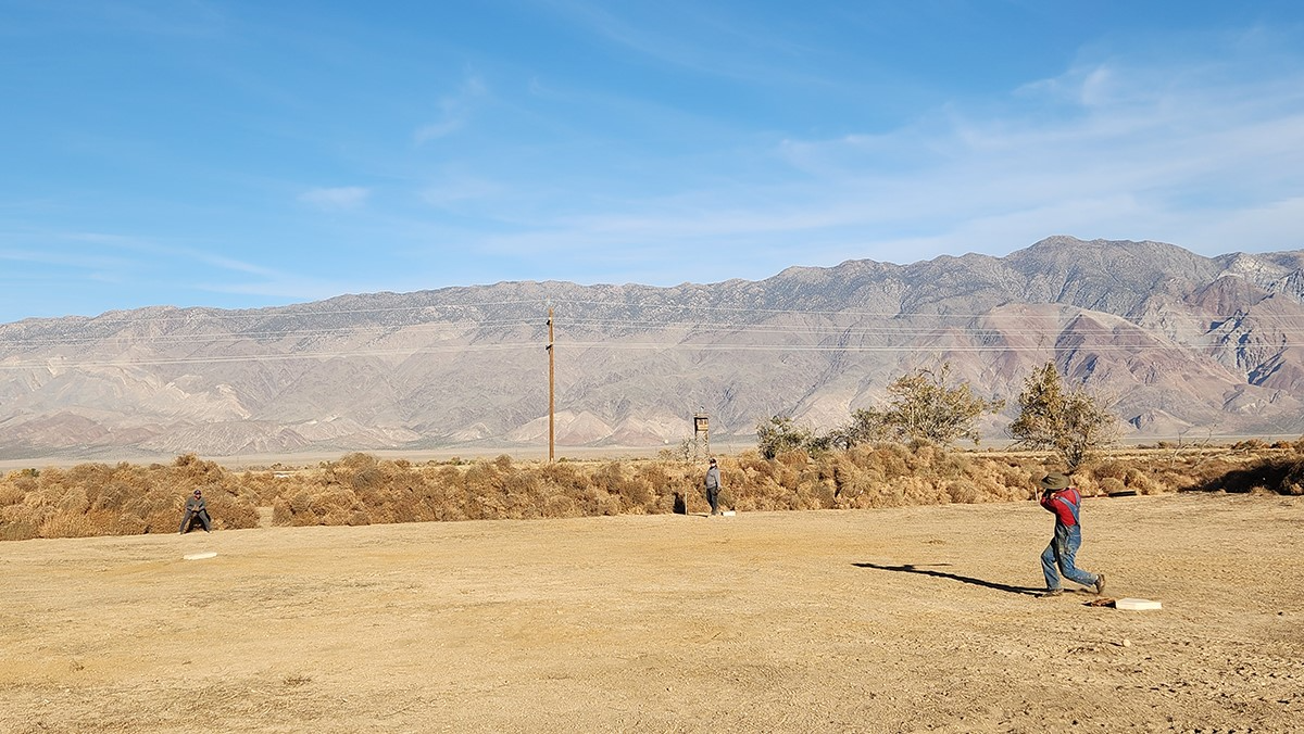 Volunteers played another game of baseball after clearing the infield and placing bases on the field at Manzanar. group plays baseball on dirt field surrounded by bushes and mountains