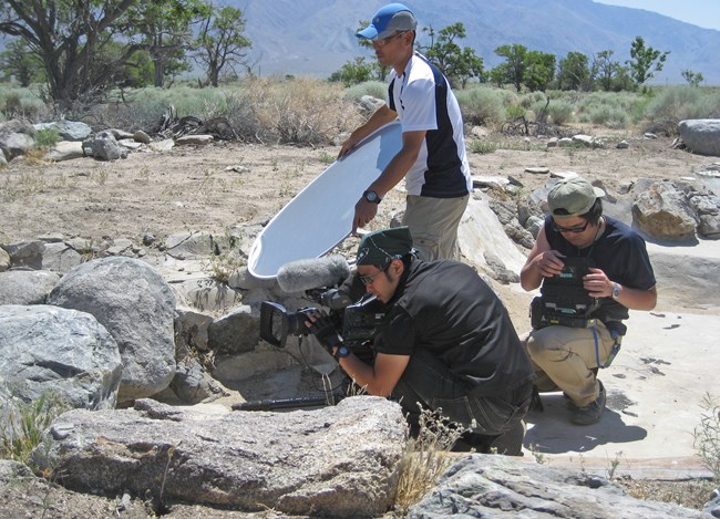 Three men with camera equipment crouch in an empty cement pond surrounded by rocks and desert