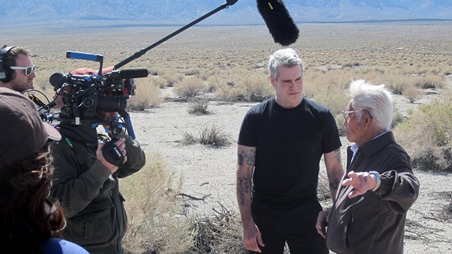 Two men stand under a boom microphone to the right of a large camera