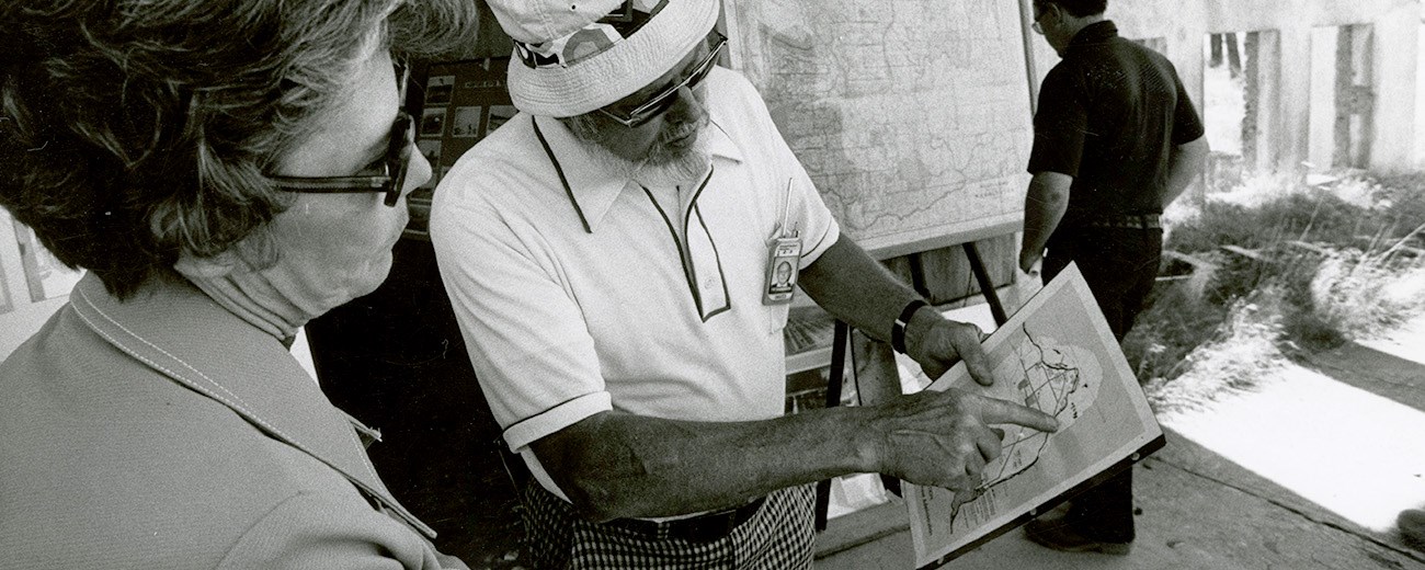Black and white photo of a man showing a woman a map. Another man looks at a large map in the background.