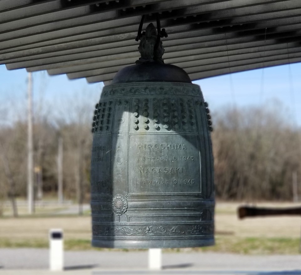 Large gray bell hangs from wooden structure above with a park in the background.