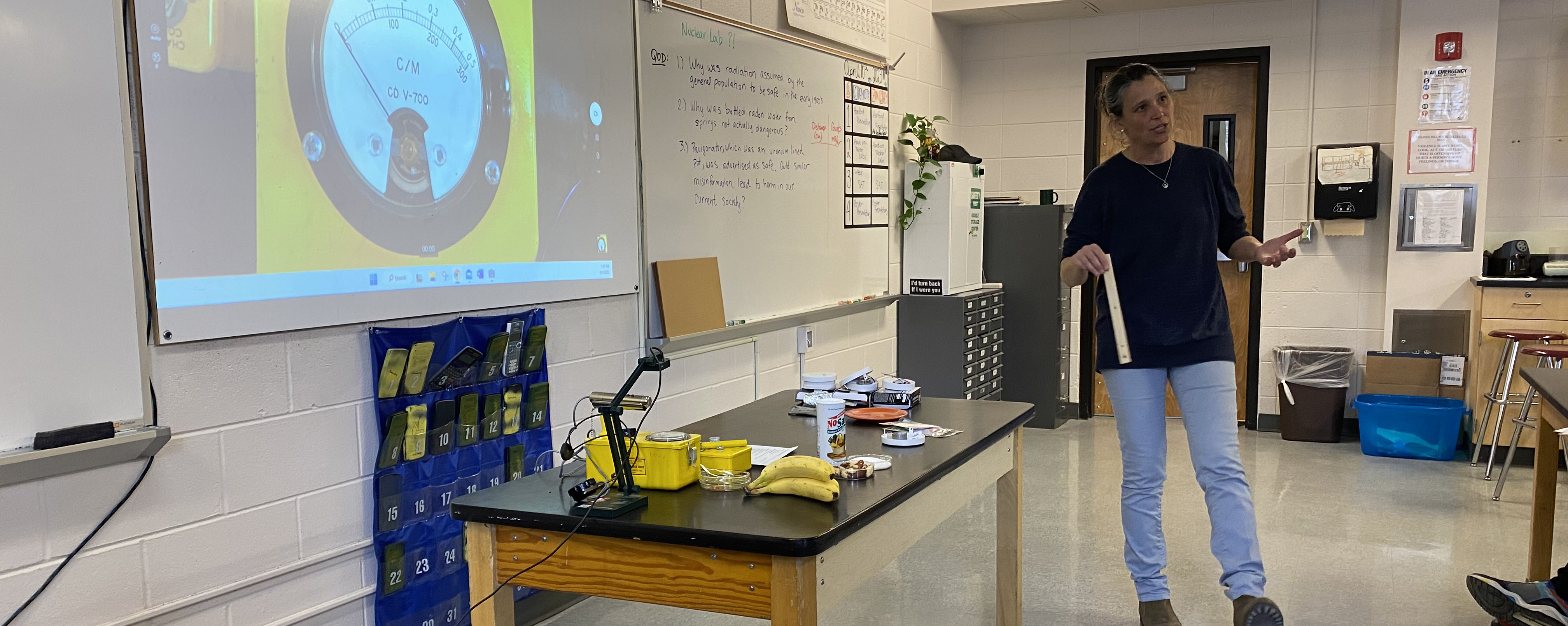 Woman stands near a table with scientific supplies and speaks to a class.