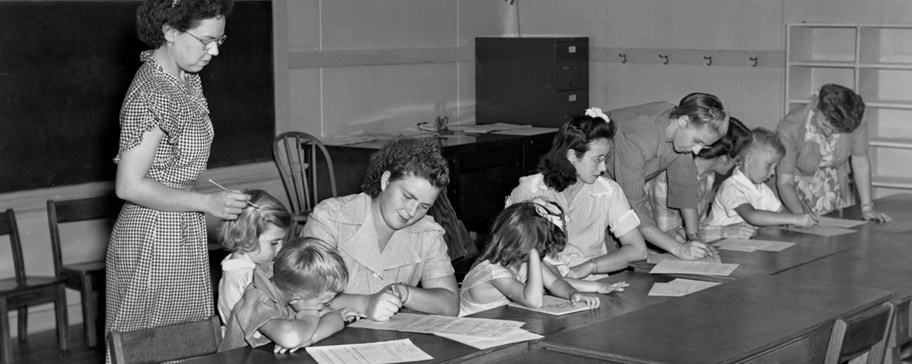Four adults and 5 children sit at desks working on papers. One teacher stands and looks over two of the seated students.