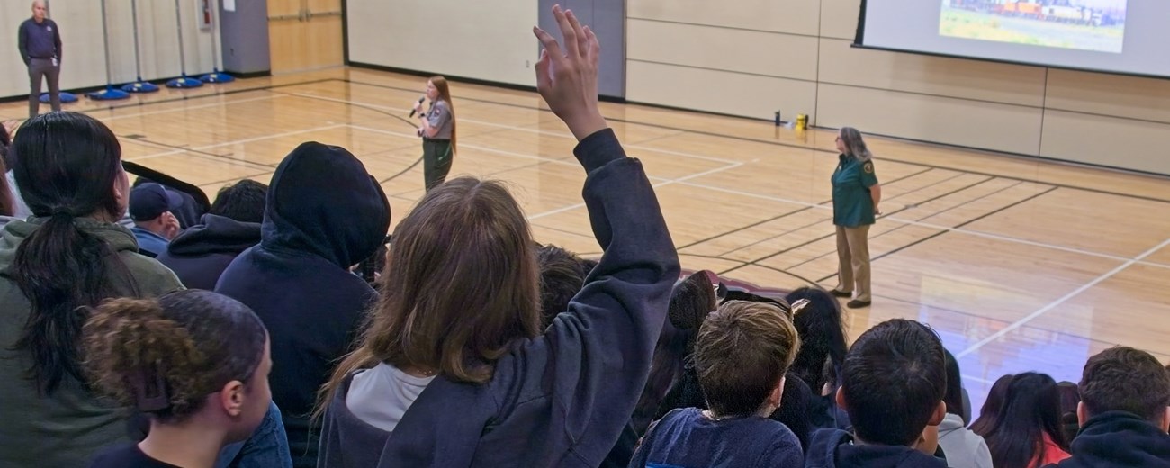 The backs of students sit on bleachers over a basketball court with two uniformed women speaking.