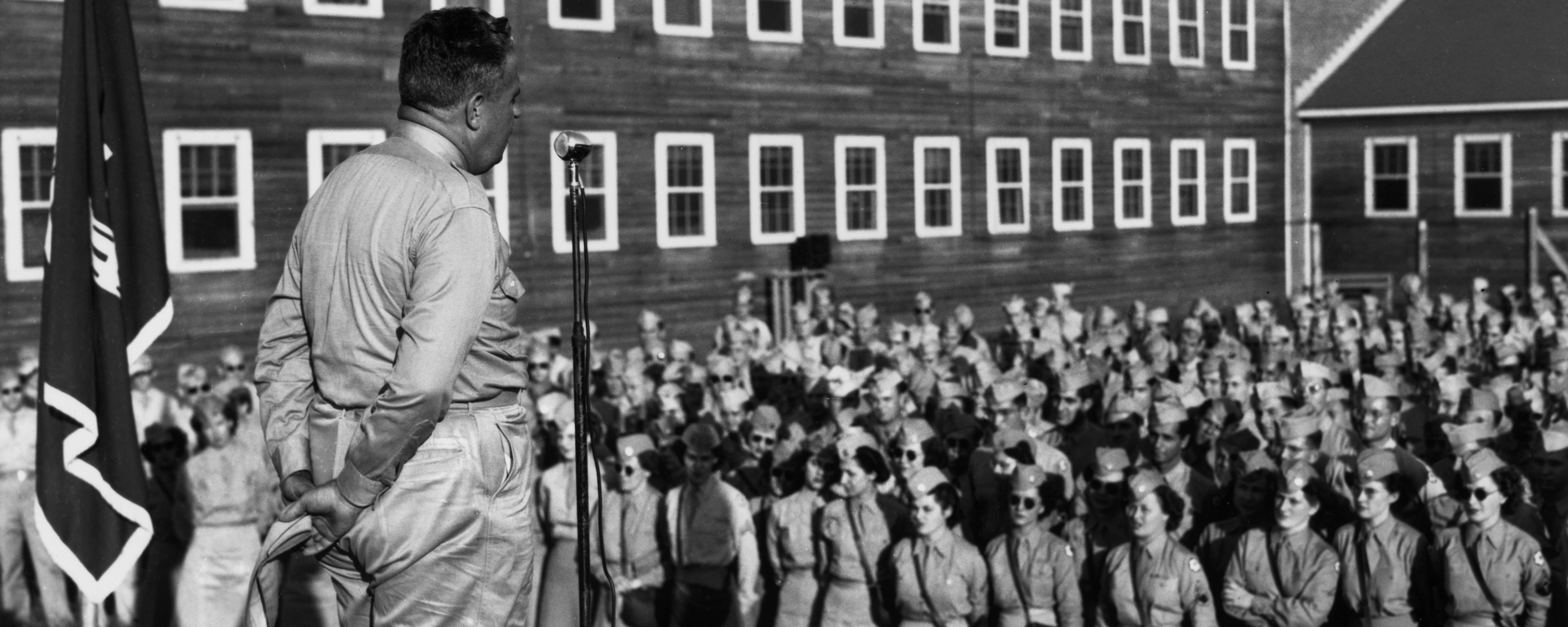 A man speaks into a microphone while facing a crowd of about 100 people wearing military uniforms listening to him.