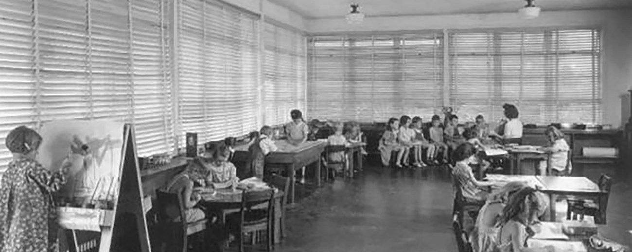 A classroom scene from 1944. 1 child stands at easel painting; groups of children sit at four tables; a teacher reads to a group of six in the back of the room.