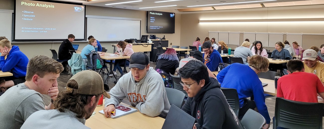 30 students sit in groups at tables with a display that says “Photo Analysis” on the board.