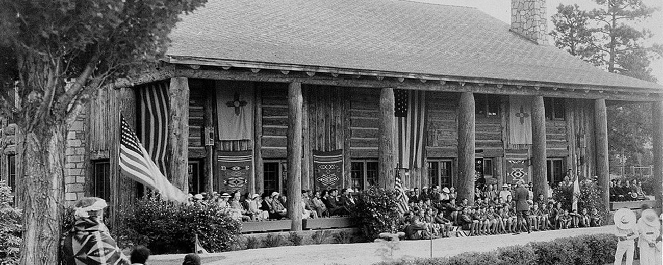 Black and white photo of a large wooden lodge with dozens of people on the porch and in the yard.