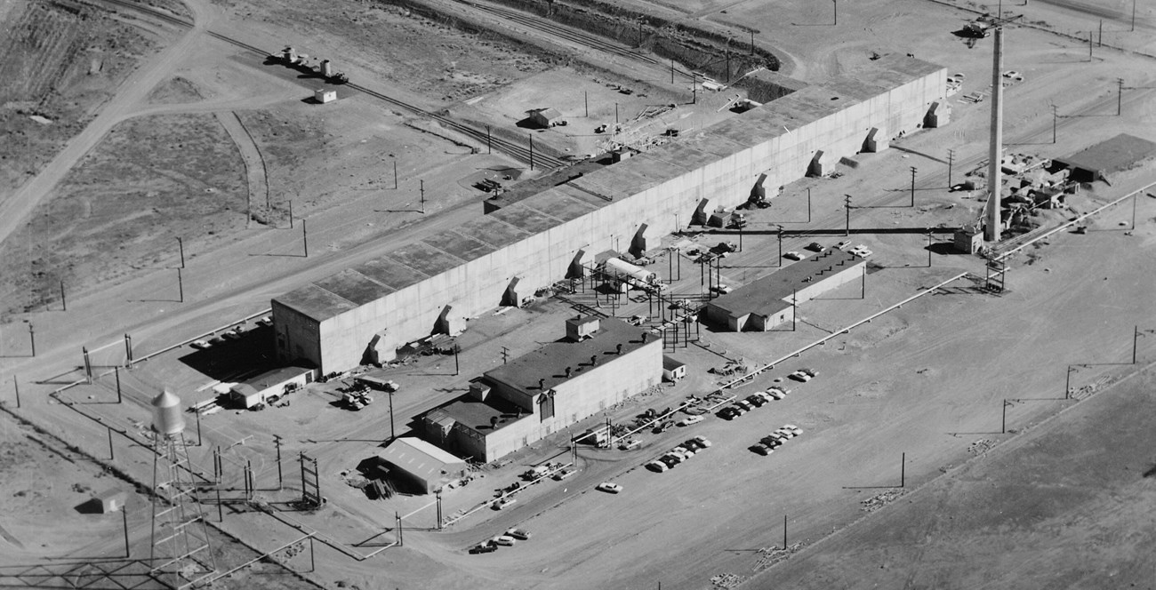 Aerial view of a long slender industrial concrete building with tall ventilation stack in a barren landscape.