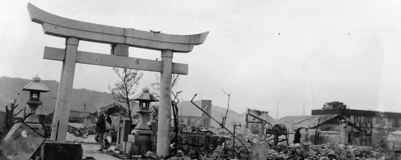 A black and white image show a man walking under Japanese architecture surrounded by destruction.