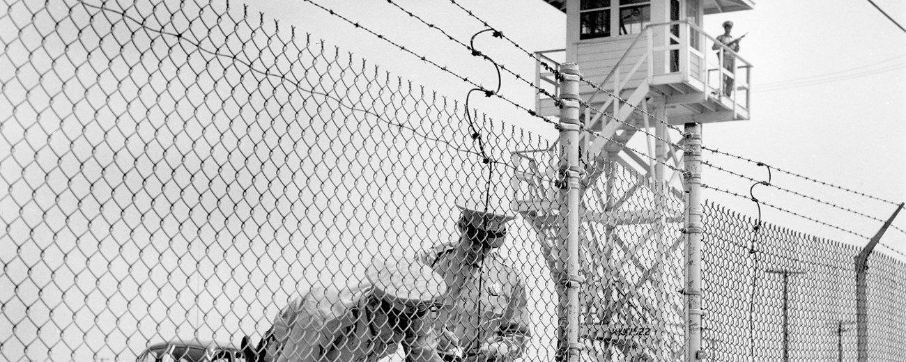 A black and white photo of two men in uniforms behind a barbed-wire fence. In the background there is a guard in the tower.