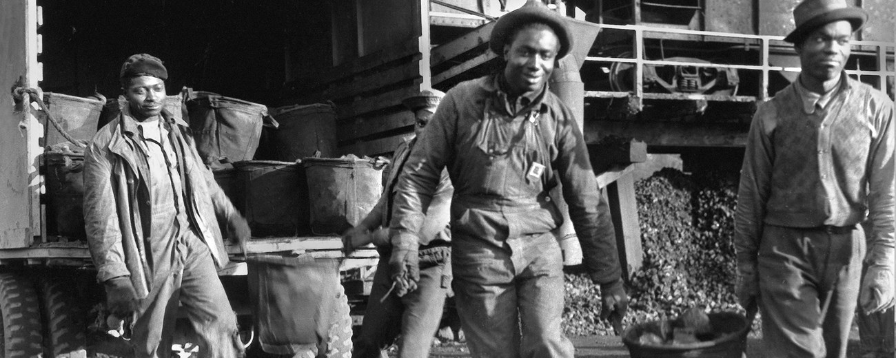 A Black and white photo of three men by a train and near a pile of what looks like coal. Two of the men are carrying a bucket.