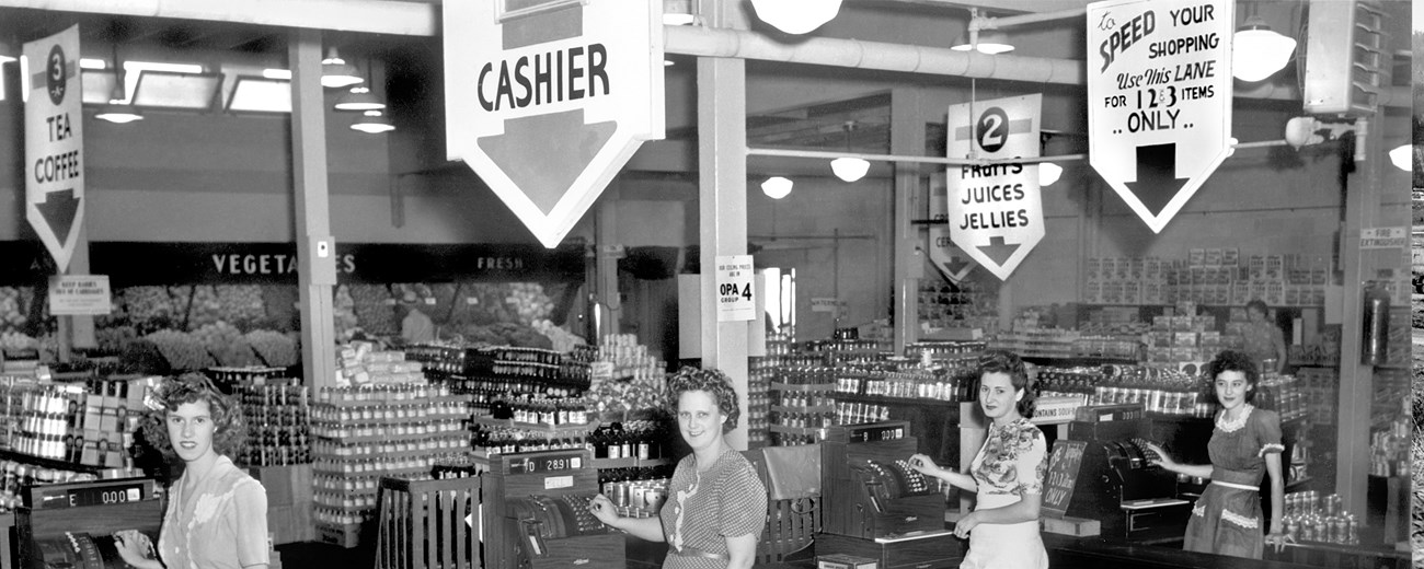 A black and white photo of several woman standing in lanes with items of food in aisles behind them.