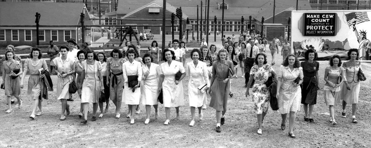 Black and white photo of tens of women, some dressed in white uniforms, walking toward the camera.  Behind them are some buildings.