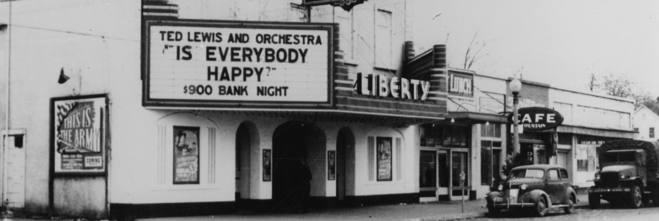 A street in a town with a theater with a sign that reads "Ted Lewis and Orchestra Is Everybody Happy?"