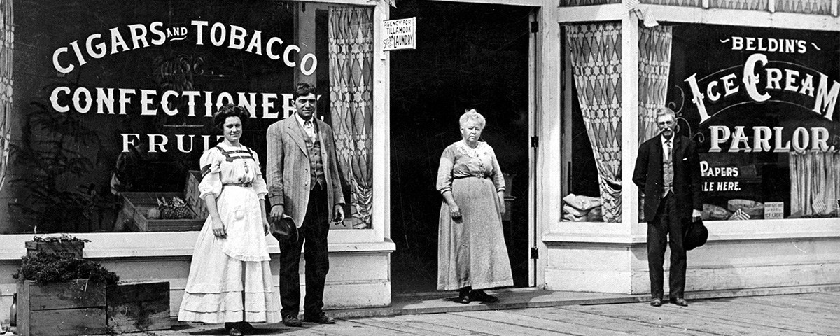 A black and white photo of two men and two women standing in front of a store. The printed letters on the storefront windows advertise cigars, fruit, and ice cream.