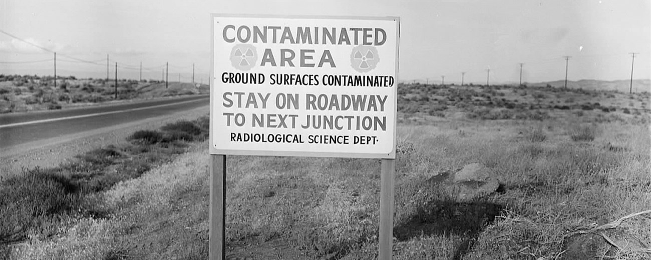A sign on a desert road reads “Contaminated Area. Ground Surfaces Contaminated. Stay on Roadway to Next Junction. Radiological Science Dept.”