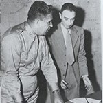 A black and white photo of General Leslie Groves and J. Robert Oppenheimer standing behind a desk, looking at paperwork.
