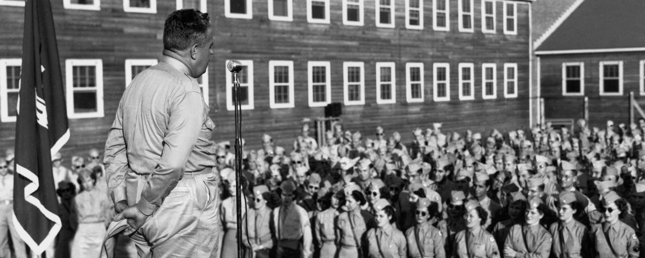 A uniformed man speaks at a microphone to several dozen people in uniform.
