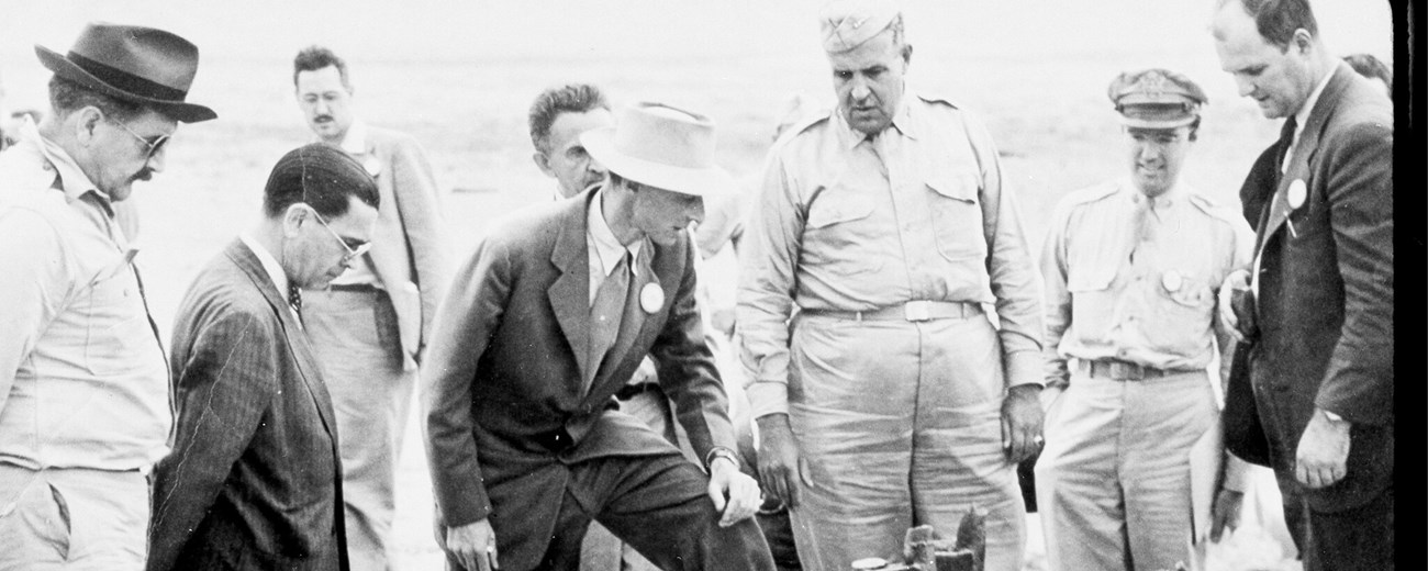 Several men, some in suits and some in uniform, gather in the desert looking at the ground.