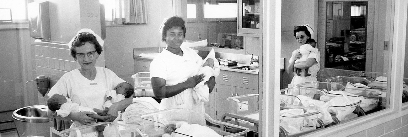 Three woman in nursing uniforms hold babies in a hospital nursery.