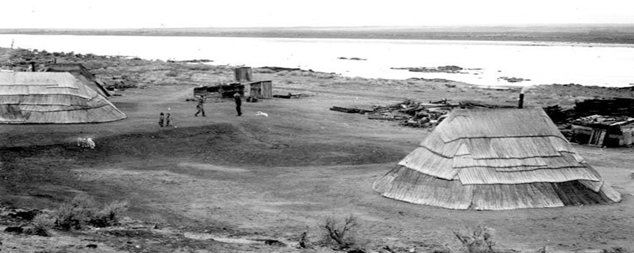Black and white photo of buildings made out of reeds by a river.
